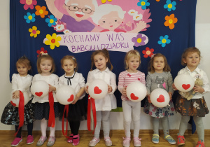 Siedem dziewczynek pozuje do zdjęcia na tle tablicy z obrazkiem przedstawiającym babcię i dziadka oraz napis Kochamy Was Babciu i Dziadku.
