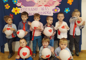 Dziewięciu chłopców pozuje do zdjęcia na tle tablicy z obrazkiem przedstawiającym babcię i dziadka oraz napis Kochamy Was Babciu i Dziadku.
