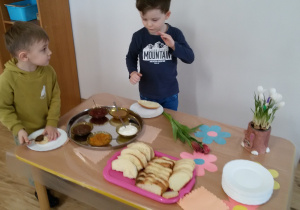 Widok na stół, na którym stoją miseczki z dżemem brzoskwiniowym, truskawkowy, twarożkiem, miodem, nutellą oraz taca z rogalami, chałką i angielką. Maciuś i Marcelek przygotowują dla siebie kanapki.