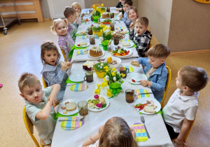Widok na dzieci siedzące przy pięknie udekorowanym wielkanocnym stole, które wspólnie spożywają przygotowane świąteczne śniadanie.