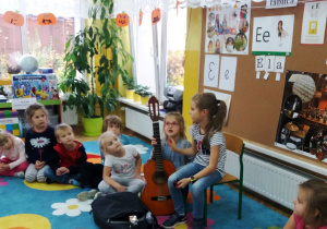 Dzieci siedzą w kręgu na dywanie, pod tablicą do zajęć na krzesełku siedzi Alicja, która prezentuje dzieciom swoją gitarę.