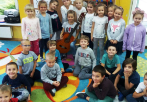 Zdjęcie grupowe. Alicja siedzi na krześle, przed sobą trzyma gitarę, wokół niej zgromadzone są dzieci. Dziewczynki stoją, a chłopcy siedzą na dywanie.