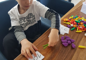 Julek układa plastikowe warzywa i znak matematyczny z plastikowych patyczków.