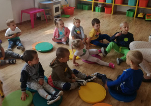Grupa dzieci siedzi na kolorowych krążkach, przed nimi na krześle siedzi nauczycielka, która czyta im bajkę.
