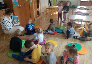 Na pierwszym planie widać siedzące na kolorowych krążkach dzieci, które patrzą na obrazek w czytanej przez nauczycielkę książce.