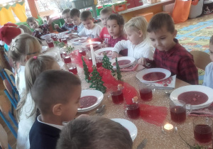 Dzieci w swiątecznych strojach siedzą przy stole. Na stole jest wigilijny obiad i świąteczne dekoracje.