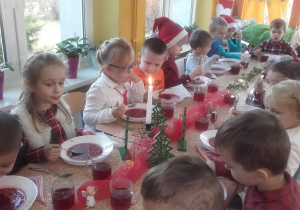 Dzieci w swiątecznych strojach siedzą przy stole. Na stole jest wigilijny obiad i świąteczne dekoracje.