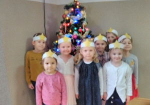 Dziewczynki stojąc przy choince pozują do zdjęcia. Mają na głowie opaski z gwiazdą.