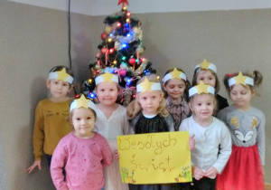 Dziewczynki stojąc przy choince pozują do zdjęcia. Mają na głowie opaski z gwiazdą. Dziewczynka stojąca w środku grupy trzyma kartkę z napisem Wesołych Świąt.