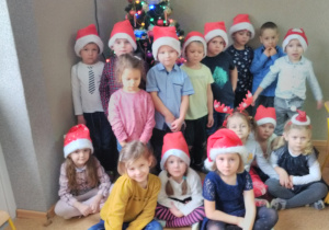 Grupa dziewczynek i chłopców stojących przy choince. Mają na głowach czerwone mikołajkowe czapeczki.