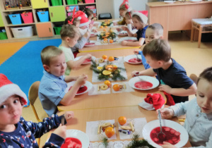 Dzieci siedzące przy stole udekorowanym stroikami ze świerku, mandarynkami i złotymi bombkami. Na stole wigilijne potrawy: czerwony barszczyk z kluskami, rybkę, kompot