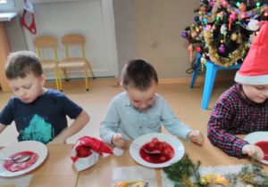 Dzieci siedzące przy stole udekorowanym stroikami ze świerku, mandarynkami i złotymi bombkami. Na stole wigilijne potrawy: czerwony barszczyk z kluskami, rybkę, kompot