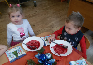 Zuzia i Mikołaj siedzą przy wigilijnym stole i jedzą barszczyk czerwony z kluseczkami. Na stole leżą serwetki, dekoracje i stroiki świąteczne.