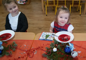 Laura i Marysia siedzą przy wigilijnym stole i jedzą barszczyk czerwony z kluseczkami. Na stole leżą serwetki, dekoracje i stroiki świąteczne.