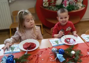 Marysia i Fabian siedzą przy wigilijnym stole i jedzą barszczyk czerwony z kluseczkami. Na stole leżą serwetki, dekoracje i stroiki świąteczne.
