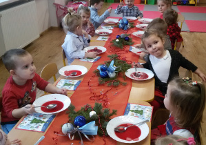 Maluszki siedzą przy wigilijnym stole i jedzą barszczyk czerwony z kluseczkami. Na stole leżą serwetki, dekoracje i stroiki świąteczne.