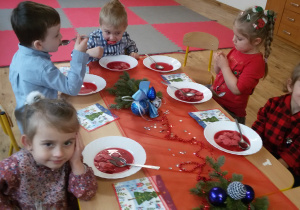 Maluszki siedzą przy wigilijnym stole i jedzą barszczyk czerwony z kluseczkami. Na stole leżą serwetki, dekoracje i stroiki świąteczne.