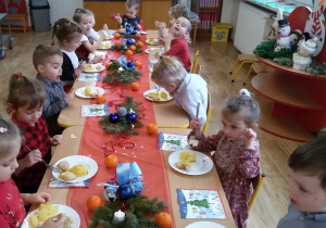 Maluszki siedzą przy wigilijnym stole i jedzą rybę z ziemniakami i kapustą. Na stole leżą serwetki, dekoracje i stroiki świąteczne.