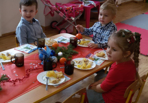 Nela, Maciuś i Olek siedzą przy wigilijnym stole i jedzą rybę z ziemniakami i kapustą. Na stole stoją kubki z kompotem oraz leżą serwetki, dekoracje i stroiki świąteczne.