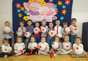 Maluszki pozują do zdjęcia grupowego trzymając w rękach białe baloniki z czerwonym serduszkiem. W tle obrazek przedstawiający babcię i dziadka oraz napis Kochamy Was Babciu i Dziadku.