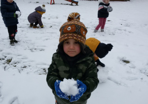 Widok na zaśnieżony ogród przedszkolny i dzieci, które lepią ze śniegu kule. Na pierwszym planie stoi Olek prezentując swoją kulę śniegową.