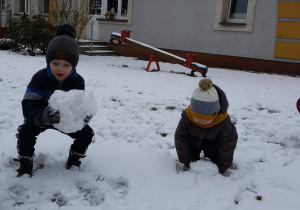 Widok na zaśnieżony ogród przedszkolny i dzieci, które lepią i toczą śnieżne kule.