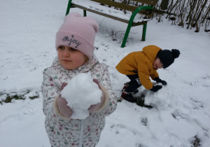 Widok na zaśnieżony ogród przedszkolny. Nela i Adaś lepią śnieżne kule.