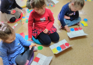 Dziewczynki siedząc na podłodze układają na planszy w kratkę figury geometryczne płaskie według kodu (czerwone koło, czerwony kwadrat, niebieskie koło, żółty kwadrat).