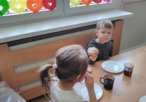 Dzieci siedząc przy stoliku spożywają lukrowane pączki.