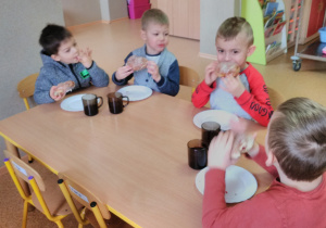 Dzieci siedząc przy stoliku spożywają lukrowane pączki.