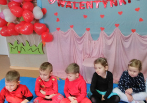 Dzieci siedzące na dywanie obok siebie i przekazujące sobie z rąk do rak „pluszowe serduszko”. W tle czerwone, różowe i białe balony w kształcie serca oraz napis „Walentynki”.