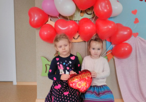 Dwie dziewczynki pozują do zdjęcia trzymając w dłoniach „pluszowe serce”. W tle dekoracja z balonów w kształcie serca, w kolorze czerwonym, różowym i białym.