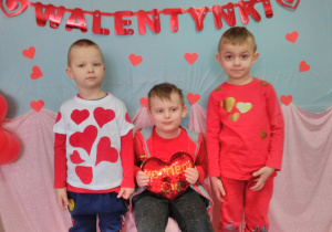 Trzech chłopców w strojach czerwonych pozujących do zdjęcia. W tle czerwony napis „Walentynki”.