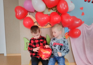 Dwóch chłopców pozuje do zdjęcia trzymając w dłoniach „pluszowe serce”. W tle dekoracja z balonów w kształcie serca, w kolorze czerwonym, różowym i białym.