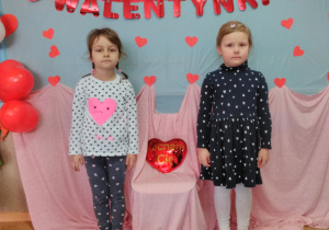 Widok na dwie dziewczynki, które pozują do zdjęcia. Między nimi na krześle „pluszowe serce” W tle czerwony napis „Walentynki”.