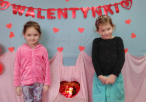 Dwie dziewczynki stojąc pozują do zdjęcia. Między nimi na krześle „pluszowe serce” W tle czerwony napis „Walentynki”.