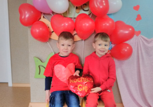 Widok na dwóch chłopców pozujących do zdjęcia, którzy trzymają w dłoniach „pluszowe serce”. W tle dekoracja z balonów w kształcie serca, w kolorze czerwonym, różowym i białym.
