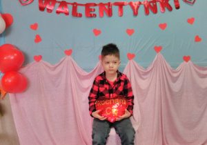 Chłopiec siedzący na krześle i trzymający w dłoni „pluszowe serce”. W tle czerwony napis „Walentynki”.