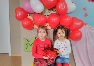 Widok na dwie dziewczynki siedzące na krześle i trzymające w dłoni „pluszowe serce”. W tle dekoracja z balonów w kształcie serca.