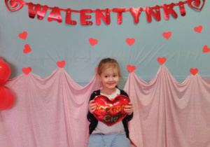 Widok na dziewczynkę siedzącą na krześle i trzymającą w dłoni „pluszowe serce”. W tle czerwony napis „Walentynki”.