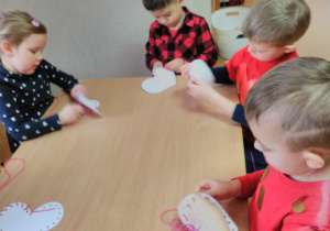 Dzieci siedzące przy stole i przeplatające przez dziurki białego serduszka różową włóczkę.