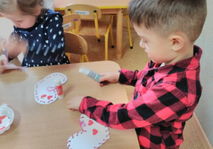Dzieci siedzące przy stole i przeplatające przez dziurki białego serduszka różową włóczkę.
