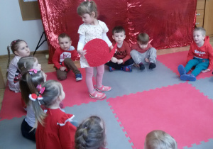 Widok na dzieci siedzące w kółeczku, w środku stoi Oliwia trzymając w rękach bibułkowe serce.
