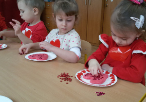 Mikołaj, Oliwia i Nadia ozdabiają swoje serca z masy solnej czerwonymi i różowymi cekinami.