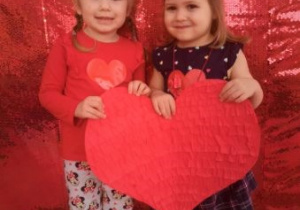 Zuzia i Lena pozują do zdjęcia z papierowym, dużym sercem. W tle czerwony, cekinowy materiał.