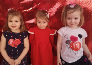 Trzy dziewczynki pozują do zdjęcia na czerwonym tle.