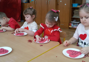 Widok na siedzące przy stoliku dzieci, które ozdabiają ulepione przez siebie serduszka różowymi i czerwonymi cekinami.
