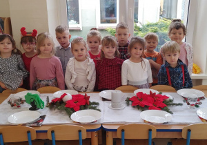 Dzieci pozują do zdjęcia w świątecznych strojach. Dzieci stoją za światecznie udekorowanym stołem.