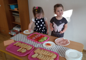 Widok na 2 dzieci, które stoją przy stoliku i przygotowują sobie kanapki. Na stoliku znajdują się tace z pieczywem, pokrojonymi warzywami oraz talerz z wędliną.