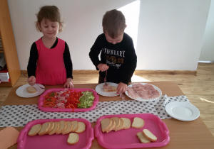 Widok na 2 dzieci, które stoją przy stoliku i przygotowują sobie kanapki. Na stoliku znajdują się tace z pieczywem, pokrojonymi warzywami oraz talerz z wędliną.